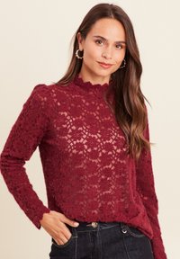 Burgunderfarbener Spitzentop mit hohem Kragen und Rüschensaum, langärmlig und mit floralem Muster. Tragen Sie dazu dunkle Jeans.