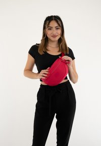 Kibővített piros deréktáska, amely szövetből készült, cipzáros záródással és lapos téglalap alakú folttal rendelkezik. A modellen fekete felső és nadrág található.