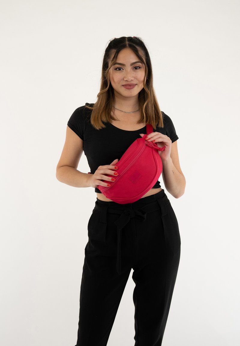 Kibővített piros deréktáska, amely szövetből készült, cipzáros záródással és lapos téglalap alakú folttal rendelkezik. A modellen fekete felső és nadrág található.