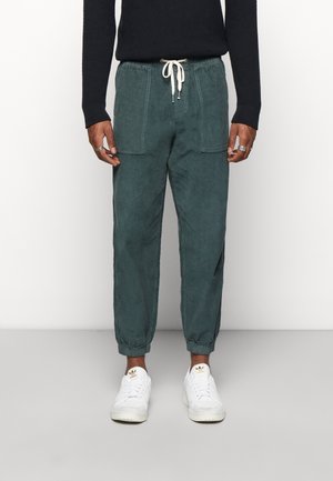 Male wearing dark green corduroy jogger pants with front pockets, white drawstring waist, black sweater, and white Adidas sneakers standing.