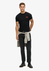 T-shirt noir à manches courtes avec logo rouge, associé à un jean noir et un pull gris noué autour de la taille. Les chaussures sont des bottes noires.