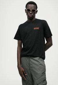 T-shirt en coton noir avec un graphique "RIPCURL" rouge et jaune. Assorti à un pantalon cargo gris. Modèle portant des lunettes de soleil claires.