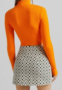 Ribbad orange topp med hög krage i mocka, kombinerad med en hög midja minikjol som har ett svart och vitt cirkulärt mönster, slät textur och figurnära silhuett.