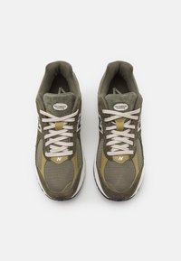 Ett par olivgröna och beige New Balance löparskor med vita snören, mesh-tyg och suedepaneler sedd från ovan.