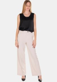 Donna con top nero senza maniche, pantaloni ampi a vita alta beige chiaro e tacchi neri a punta, in piedi davanti a uno sfondo semplice.