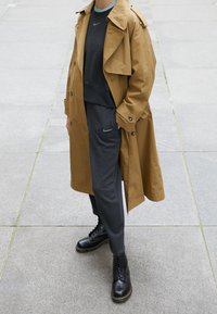 Tan trench coat over a dark gray sweatshirt and matching joggers, paired with black lace-up boots. Notable Nike logo accents on clothing.