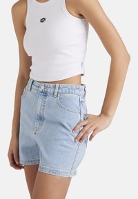 Hellblaue Jeansshorts mit hohem Bund, vorderem Knopfverschluss und zwei Seitentaschen. Kombiniert mit einem taillierten weißen Tanktop.