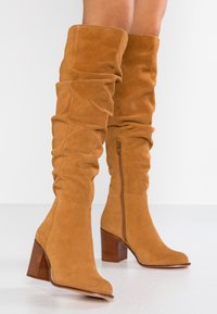 Botas de ante beige por encima de la rodilla con un diseño holgado, punta redondeada, tacón de madera grueso y detalle de cremallera lateral para facilitar su uso.