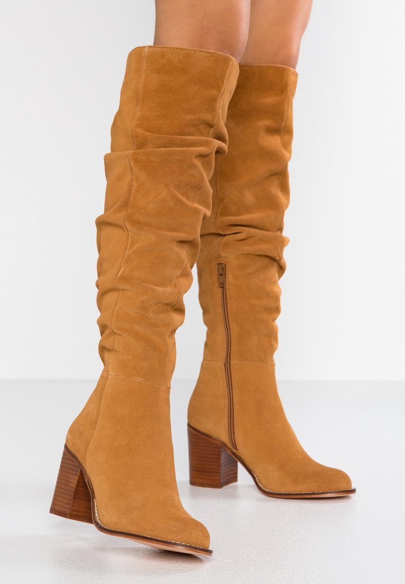 Botas de ante beige por encima de la rodilla con un diseño holgado, punta redondeada, tacón de madera grueso y detalle de cremallera lateral para facilitar su uso.