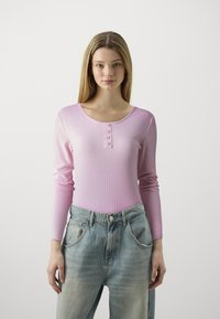 Top a maniche lunghe ribbed rosa chiaro con scollatura a barchetta e quattro bottoni decorativi, abbinato a jeans azzurri a vita alta.
