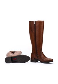 Bottes en cuir marron montant jusqu'au genou avec des fermetures éclair sur le côté, l'une se tenant debout et l'autre allongée sur sa semelle, montrant le motif de la semelle et le talon.