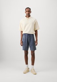 Polo shirt texturé crème avec col, associé à un short en denim délavé et des baskets beiges. Design simple, coupe décontractée, shorts à taille élastique.