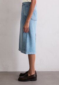 Lichte blauwe denim midi-rok met zijsplits, raw hem en achterzak, gecombineerd met donkerbruine, chunky loafers.