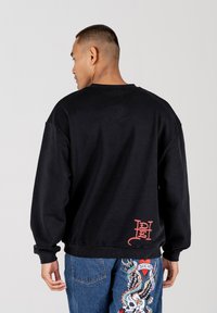 Black crewneck sweatshirt with dropped shoulders, ribbed cuffs, and a red logo at the lower back. Contrast with blue embellished jeans.