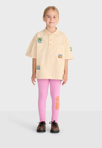 Child wearing oversized beige polo shirt with colorful patches, pink leggings with orange text on one leg, and brown shoes standing against plain background.