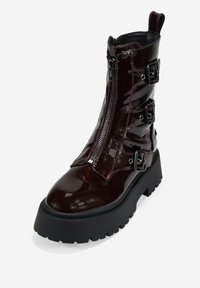 Stivale da combattimento lucido di un bordeaux scuro con suola nera spessa, zip frontale, tre fibbie laterali e linguetta sul retro.