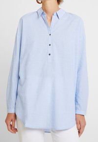 Camicia azzurro chiaro a maniche lunghe con colletto e patta a mezza bottoniera. Tessuto morbido, vestibilità rilassata e spacchi laterali all'orlo.