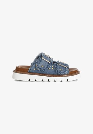 Denim-Sandalen mit fransigen Rändern, doppelten silbernen Schnallen und goldenen Nieten auf einem braunen Fußbett mit einer dicken weißen Sohle.