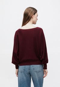 Bordowy sweter z dzianiny z szerokim dekoltem i obniżonymi ramionami. Posiada długie rękawy i prążkowany brzeg, zestawiony z jasnoniebieskimi dżinsami.