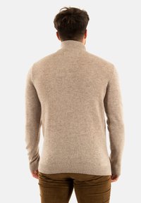 Pull en tricot beige avec un col montant, présentant une texture douce et des poignets côtelés, vu de dos, associé à un pantalon en velours côtelé marron.