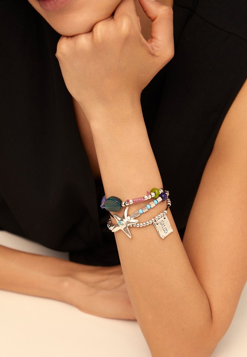 Pulsera colorida de cuentas con acentos de plata, que incluye un charm de estrella de mar y una etiqueta grabada con "SALUD"; presenta formas y texturas variadas.