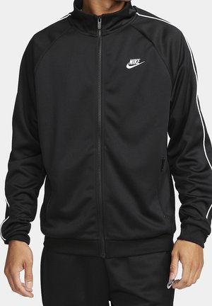 Uomo che indossa una giacca da allenamento nera Nike con strisce bianche sulle maniche e cerniera frontale, abbinata a pantaloni neri coordinati.