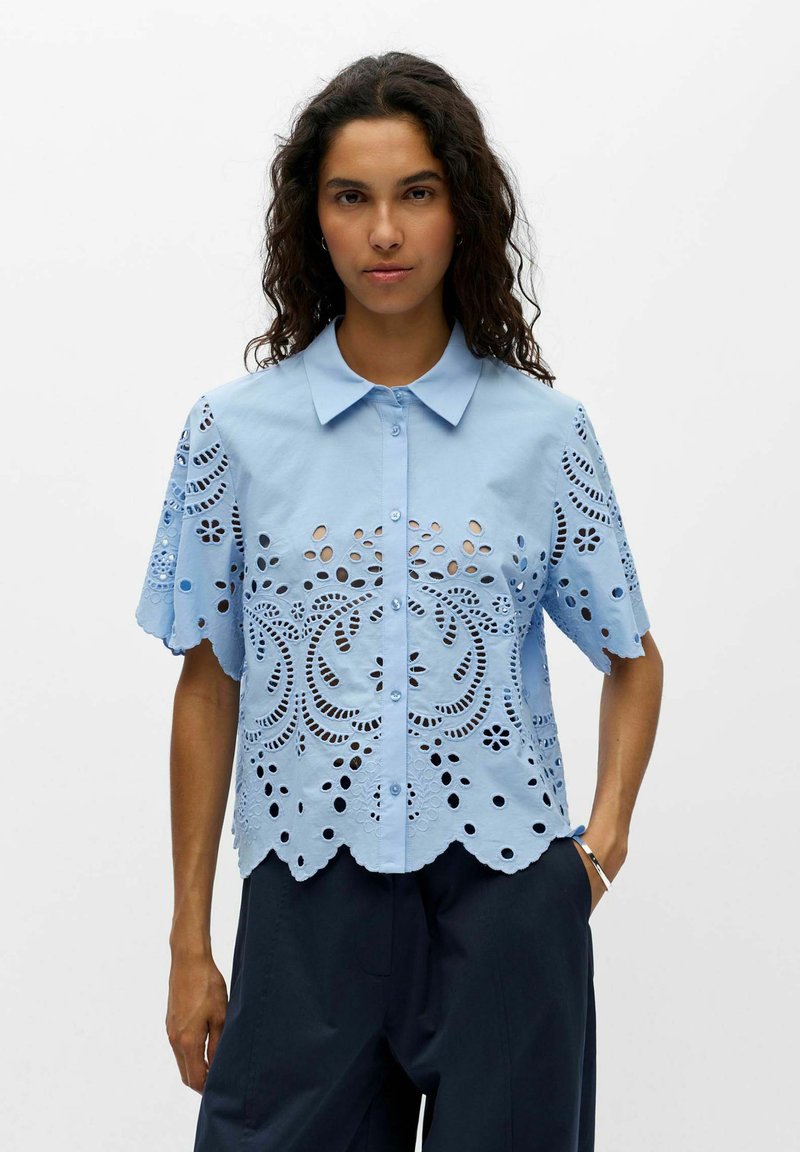 Femme aux cheveux bouclés foncés portant un chemisier bleu clair à manches courtes avec des motifs floraux découpés et un pantalon foncé sur un fond uni.