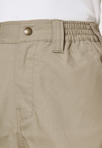 Beige werkbroeken met een elastische tailleband, sluiting met een voorste knoop, subtiel ruitpatroon en gestikte details op de zijzakken.
