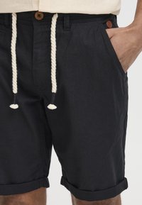 Shorts in cotone nero con finitura testurizzata, dotati di vita con cordoncino, orlo arrotolato e tasche laterali. Dettaglio con bottone marrone in vita.