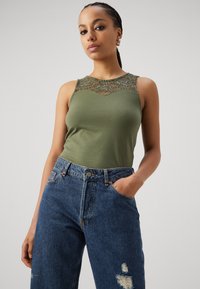 Olivenfarget tank topp med blonde detaljer i utringningen, kombinert med høyt liv, løstsittende, lett slitte blå denim jeans.
