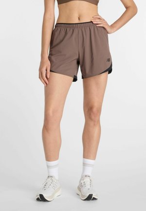 Frau trägt braune Sportshorts, weiße Socken und weiße New Balance Laufschuhe, steht mit einer Hand in der Hüfte.