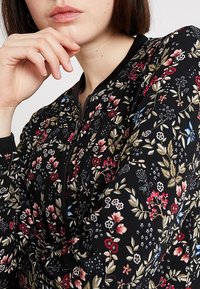 Veste bomber fleurie avec une base en tissu noir, des motifs floraux rouges et bleus vibrants, des poignets côtelés et une fermeture éclair.