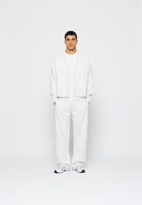 Biely bavlnený kardigán s gombíkmi, skombinovaný s bielym tričkom a zodpovedajúcimi voľnými nohavicami. Oblečenie dopĺňajú neformálne šedé tenisky.