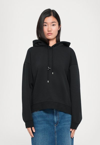 Schwarzer Kapuzenpullover mit lockerer Passform, weiten Ärmeln und Kordelzugakzenten. Kombiniert mit einem blauen Jeansrock.