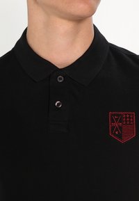 Czarna koszulka polo z klasycznym kołnierzykiem, z dwoma guzikami i czerwonym haftowanym logo w kształcie tarczy po lewej stronie klatki piersiowej. Teksturowana tkanina.