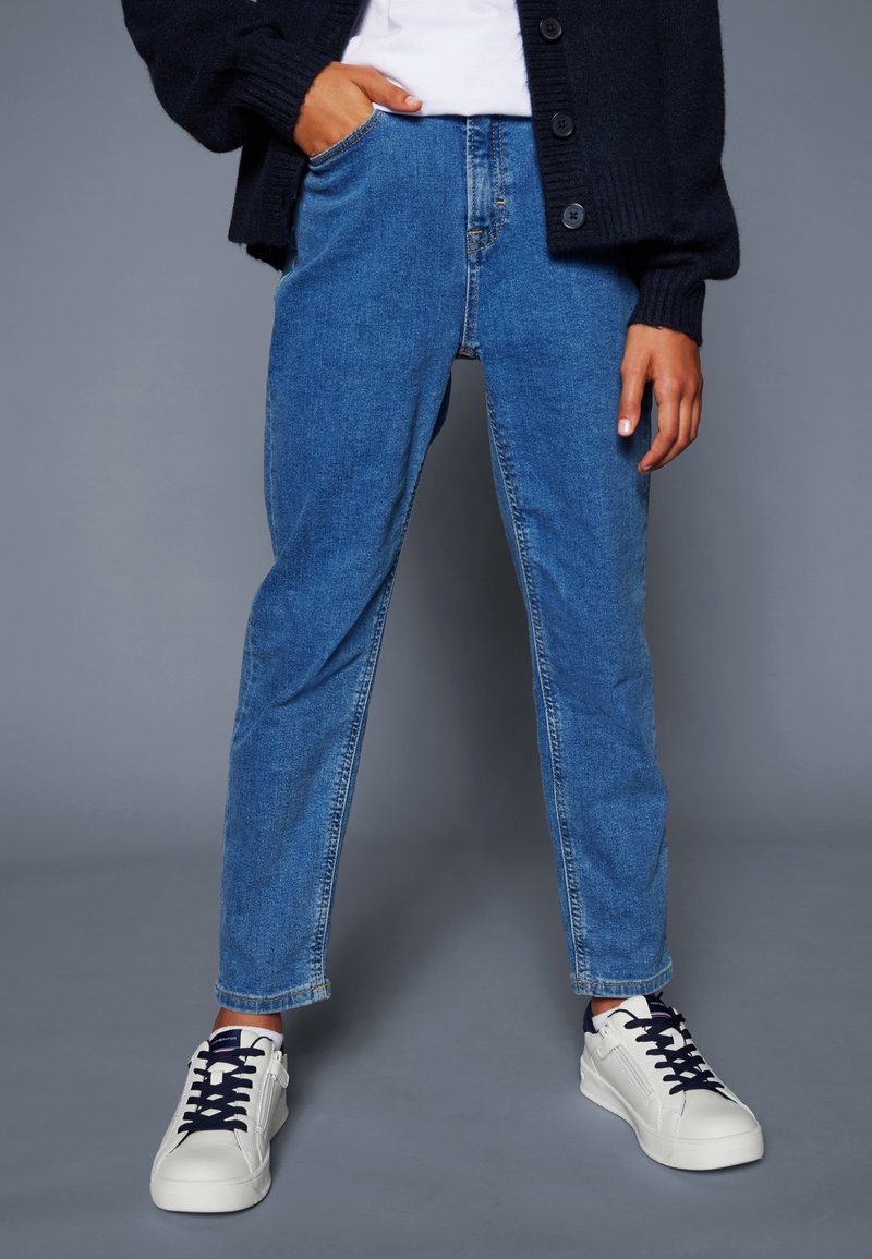 Jeans de mezclilla azules con corte recto, que presentan una cintura alta y un diseño de cinco bolsillos. Combinados con zapatillas blancas con acentos en azul marino.