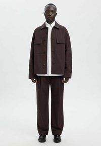 Brun jakke med to frontlommer over en hvit skjorte, kombinert med brune bukser og svarte sko. Glatt stoff med en avslappet passform.