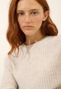 Lekki, dziany sweter z teksturowanym wzorem, okrągłym dekoltem i miękkim, neutralnym kolorem. Posiada długie rękawy i luźny krój.