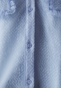 Light blue textured shirt featuring a crinkled fabric, button closure, and ruffled accents along the collar and pockets.