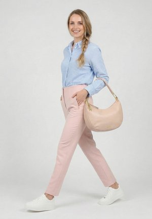 Frau geht, trägt hellblau kariertes Hemd, rosa Hose, weiße Sneaker, trägt eine beige Handtasche auf der rechten Schulter, lächelt, heller Hintergrund.