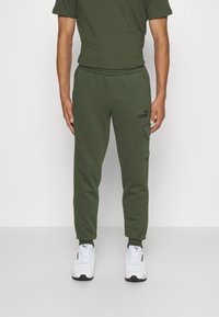 Pantalones de chándal verde oliva con cintura ajustable, bolsillo lateral tipo cargo y logo negro. Combinados con zapatillas deportivas blancas.