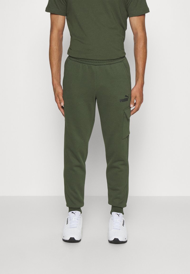 Pantalones de chándal verde oliva con cintura ajustable, bolsillo lateral tipo cargo y logo negro. Combinados con zapatillas deportivas blancas.