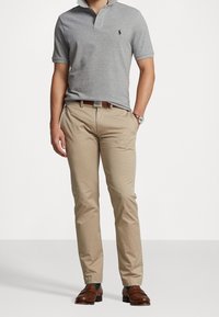 Šedé polo tričko s límečkem a malým modrým logem, kombinované s béžovými chinos kalhotami a hnědými koženými botami. Hladká, plochá látka a přiléhavý střih.