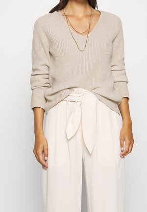 Kvinde iført beige strikket sweater med opkørte ærmer, løse cremefarvede bukser bundet i taljen og flere lag af guldkæder.