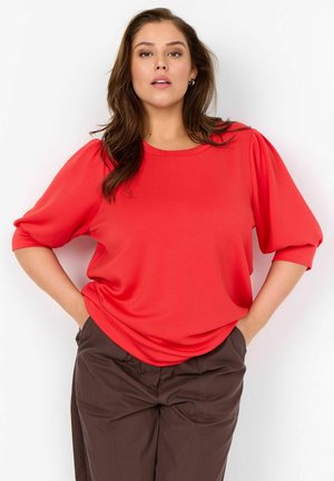 Frau mit langen braunen Haaren, die eine leuchtend rote Bluse mit Puffärmeln und dunkelbraune Hosen trägt, Hände in den Taschen, vor weißem Hintergrund.