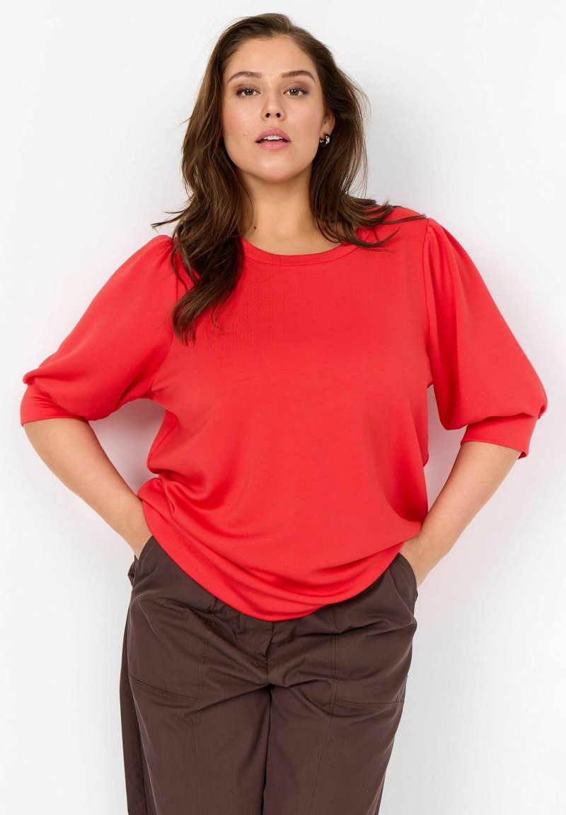 Femme aux longs cheveux bruns portant un chemisier rouge vif à manches bouffantes et un pantalon marron foncé, les mains dans les poches, sur fond blanc.