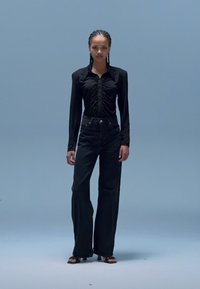 Črni top z dolgimi rokavi z nabrano detajlno obdelavo, kombiniran z visokim pasom črnih kavbojk. Model stoji na svetlo modrem ozadju.