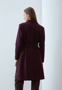 Cappotto bordeaux con vita aderente e cintura, dotato di colletto a revers e maniche lunghe, abbinato a pantaloni marrone scuro, con texture liscia.