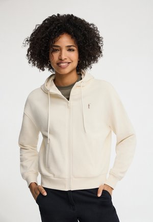 Vrouw met krullend haar die lacht, een crème kleurige hoodie met rits draagt en zwarte broek, handen in de zakken, staand tegen een effen achtergrond.