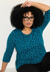 Tefarbener Langarm-Top mit schwarzem Punkte-Muster, runder Ausschnitt und weichem Stoff, kombiniert mit schwarzen Hosen.